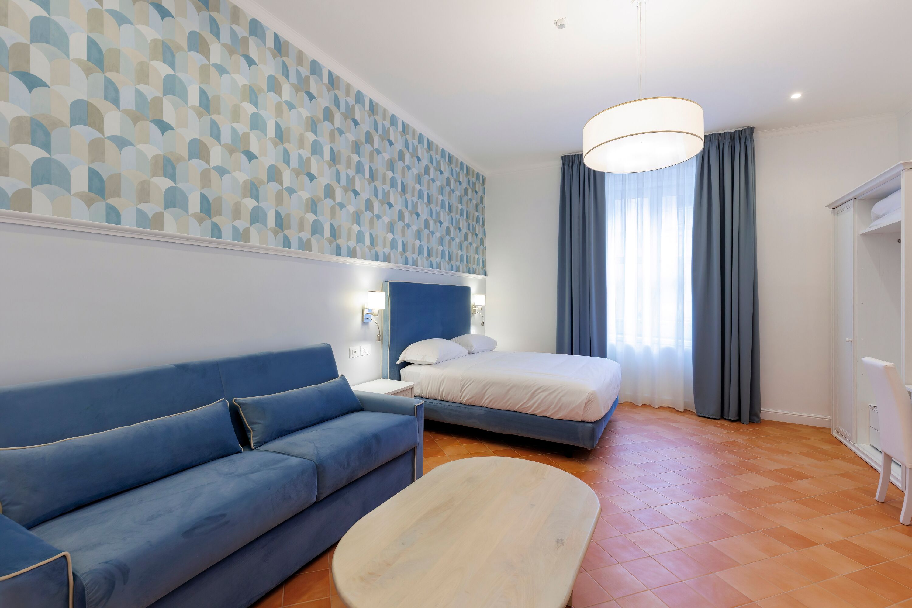 Una habitación de hotel moderna con un sofá azul, una cama con ropa de cama blanca, una mesa de centro de madera y una pared con un diseño estampado.