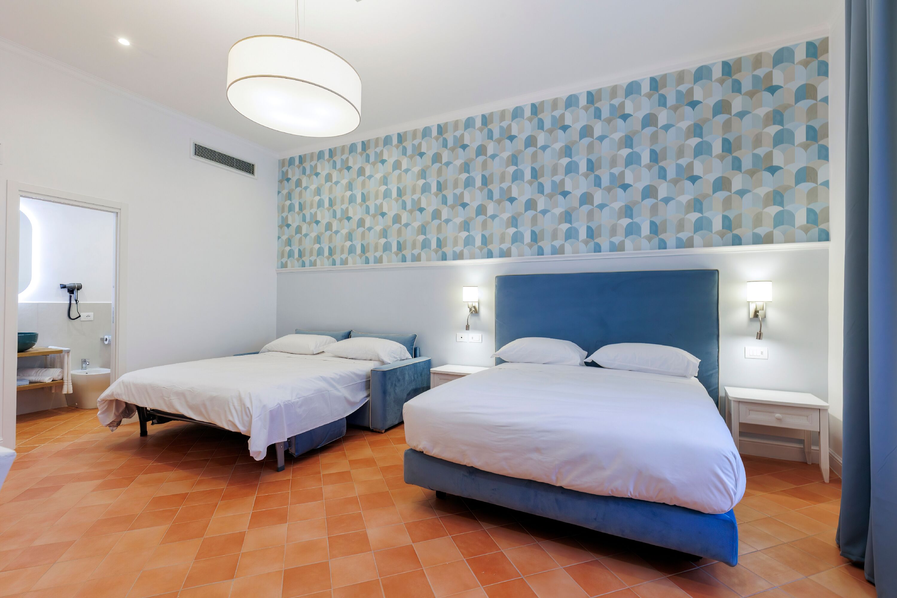 A hotel room with two beds, a blue headboard, white walls, a patterned ceiling, and a tiled floor.