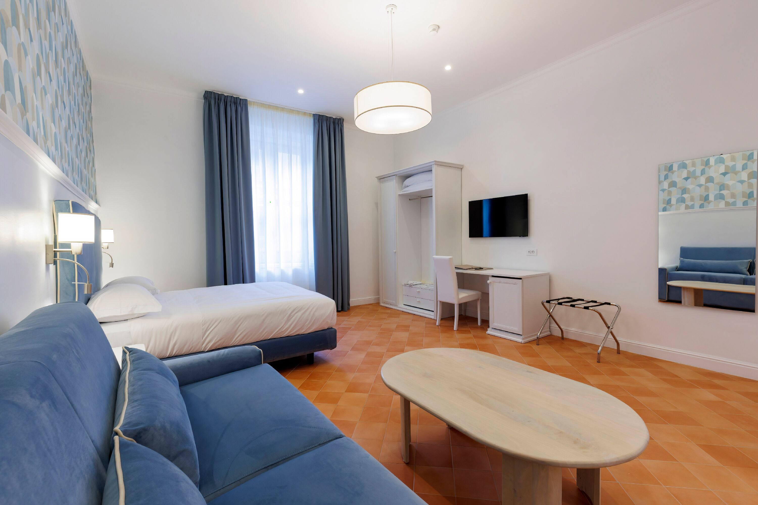 A modern hotel room with a bed, a desk, a chair, a sofa, and a TV mounted on the wall.