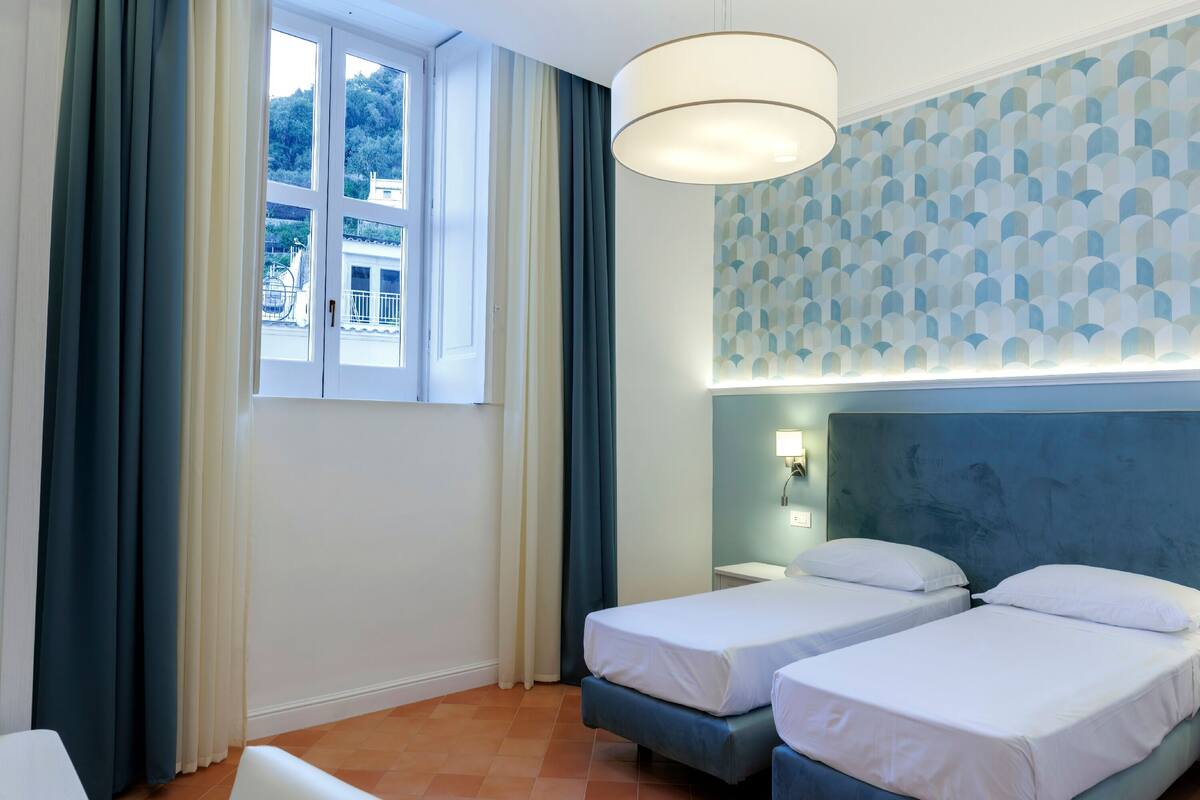 Habitación de hotel con dos camas, una ventana con cortinas, una lámpara de pared y un papel tapiz estampado.