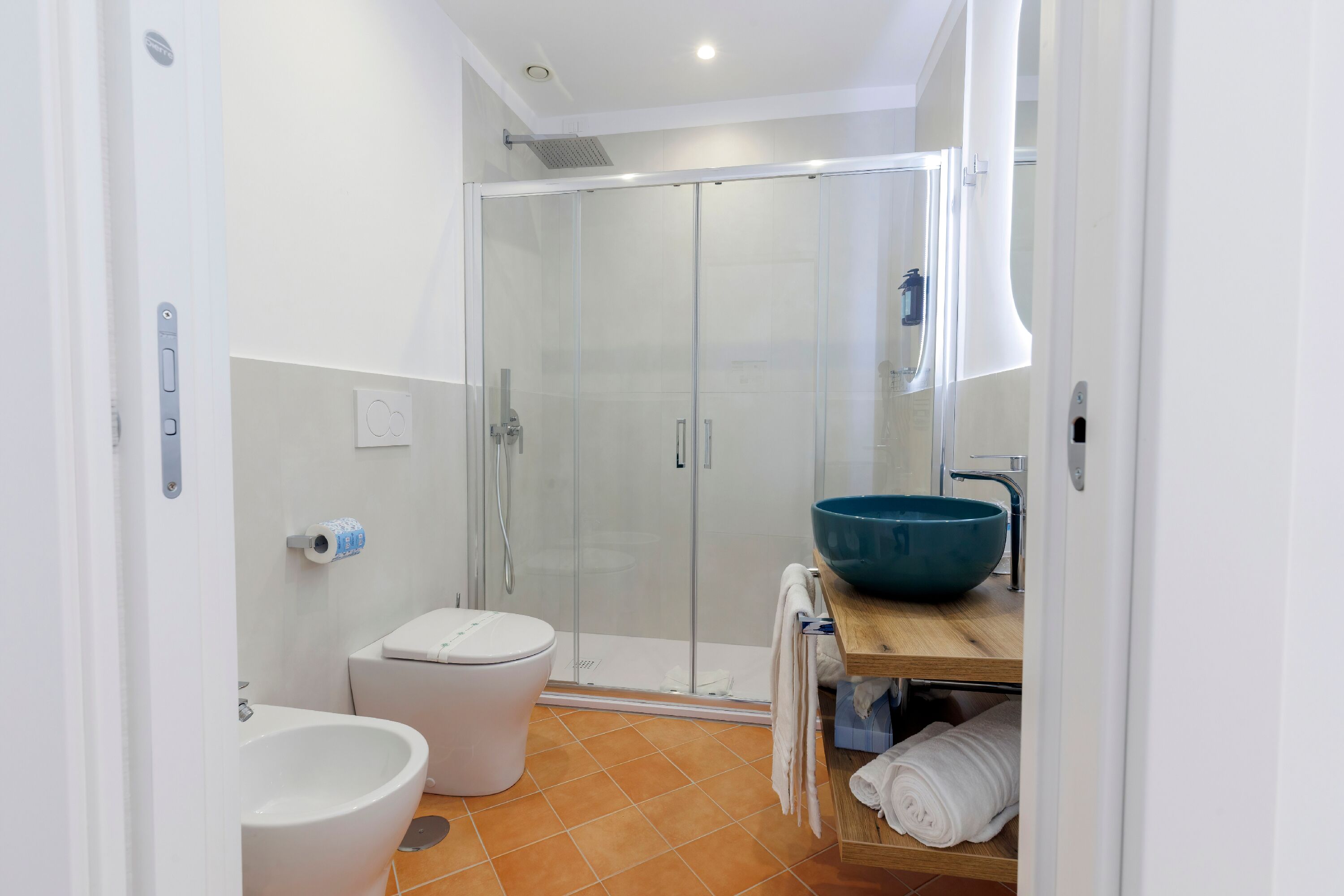 A modern bathroom with a glass shower enclosure, a white toilet, a large blue basin, and a wooden vanity.
