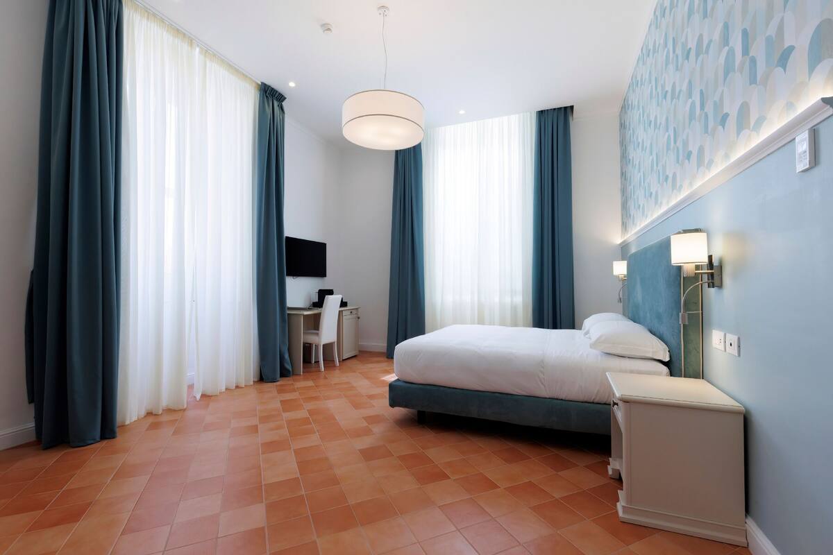 Habitación de hotel con cama, escritorio, silla, televisor y lámpara de pared.
