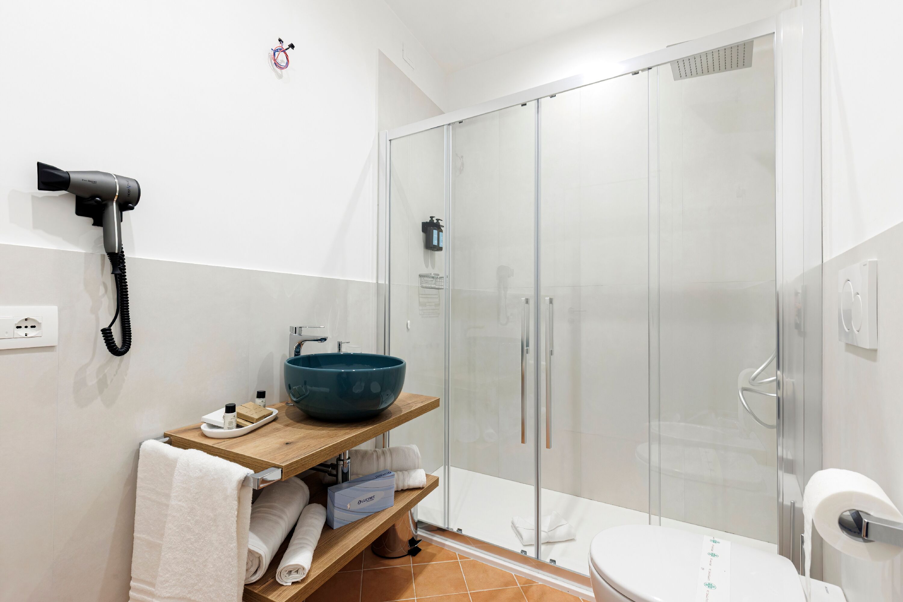 Un baño moderno con cabina de ducha de vidrio, tocador de madera con lavabo azul y secador de pelo montado en la pared.