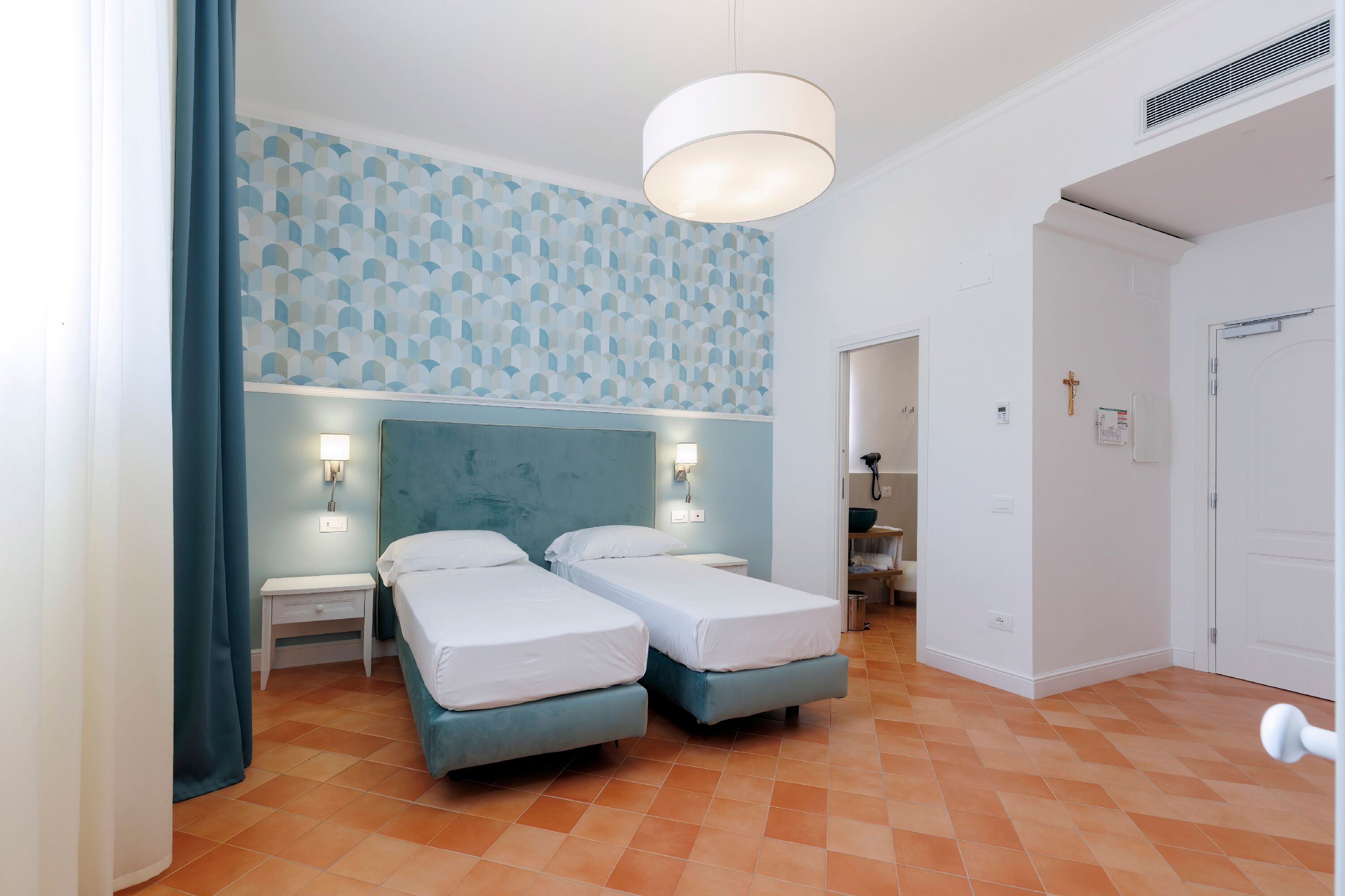 A hotel room with two beds, a blue headboard, a patterned wall, and a large window with curtains.