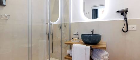 A modern bathroom with a glass-enclosed shower, a large oval mirror, a wooden vanity with a bowl sink, and a hairdryer mounted on the wall.