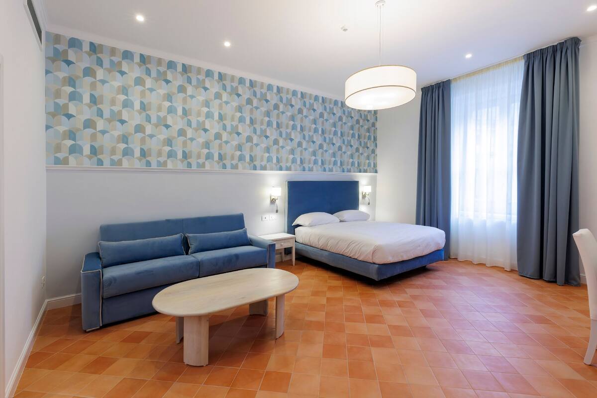 Una habitación de hotel moderna con un sofá azul, una cama, una mesita y un papel tapiz estampado.