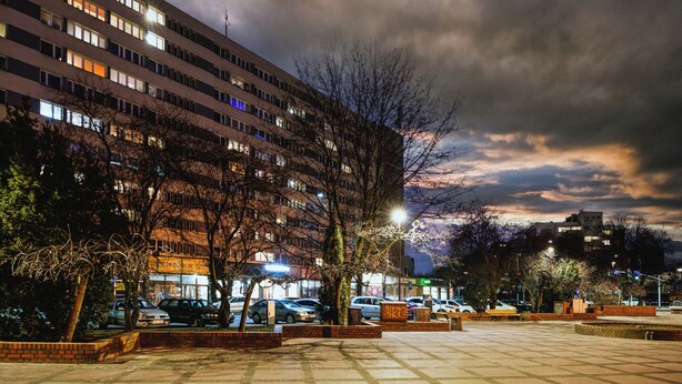 A parking lot with multiple cars, a building with lit windows, and a cloudy sky. - Amber - by Kairos Apartments (Gliwice)