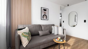 A modern living room with a grey sofa, a small round table with a plate of fruit, and a large mirror on the wall. - Amber - by Kairos Apartments (Gliwice)