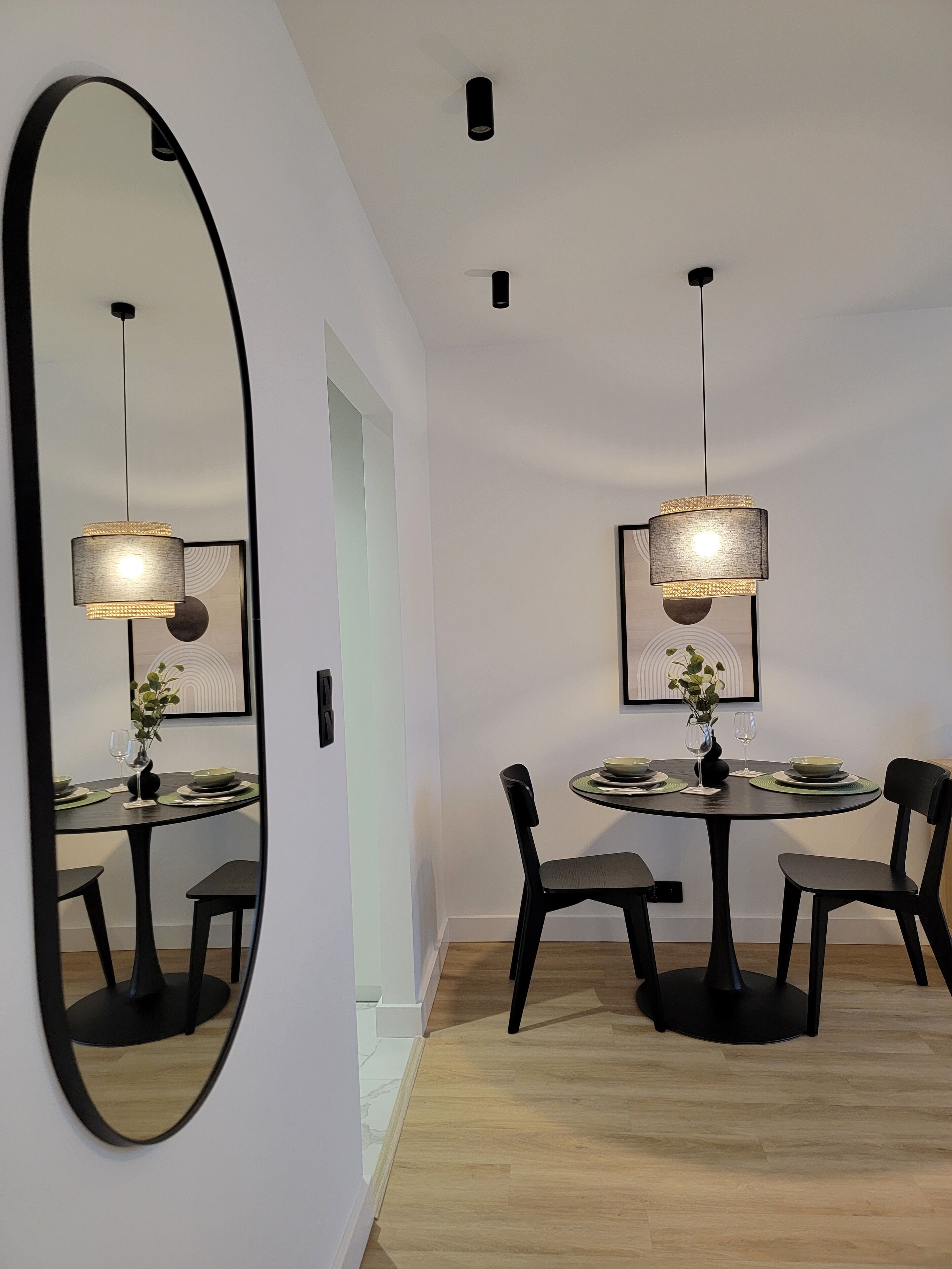 A dining area with a round table, black chairs, and two pendant lights. A large mirror is mounted on the wall to the left.