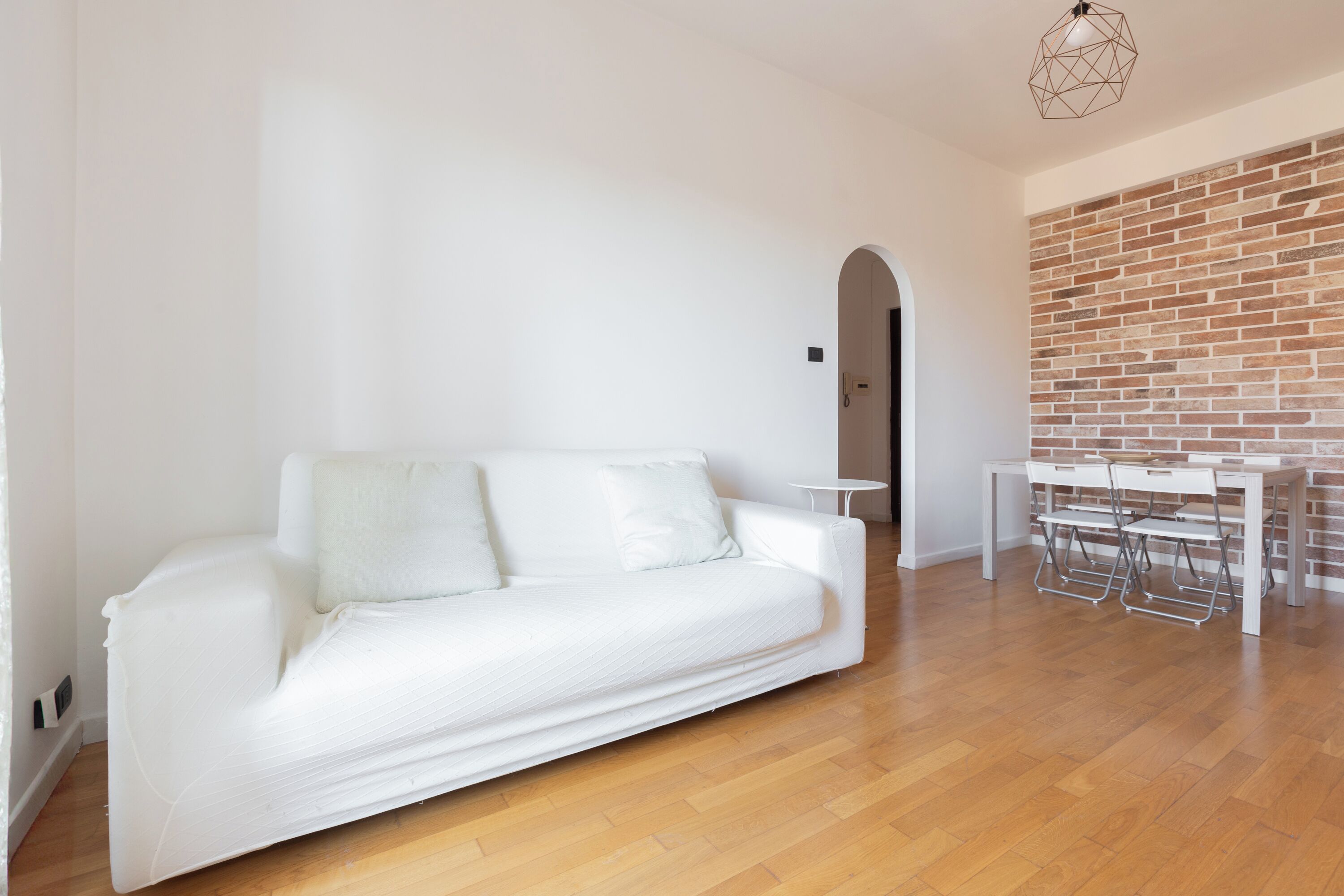 A modern living room with a white sofa, a dining table with chairs, and a brick wall.