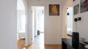 Un pasillo con una puerta blanca que da a un dormitorio, un altavoz negro sobre una mesa y un cuadro en la pared.