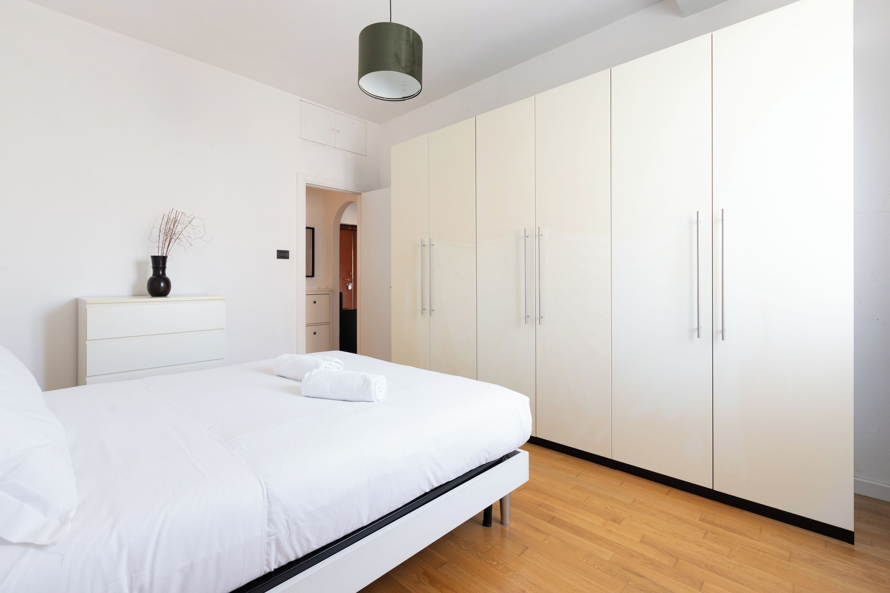 A modern bedroom with a large bed, white wardrobe, and a small bedside table with a vase.