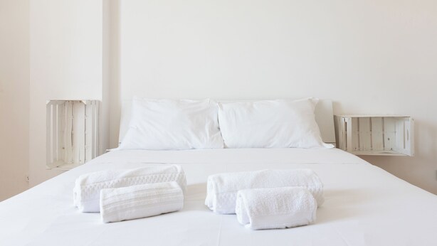 Una cama bien hecha con sábanas y almohadas blancas, dos mesitas de noche y toallas dobladas sobre la cama.