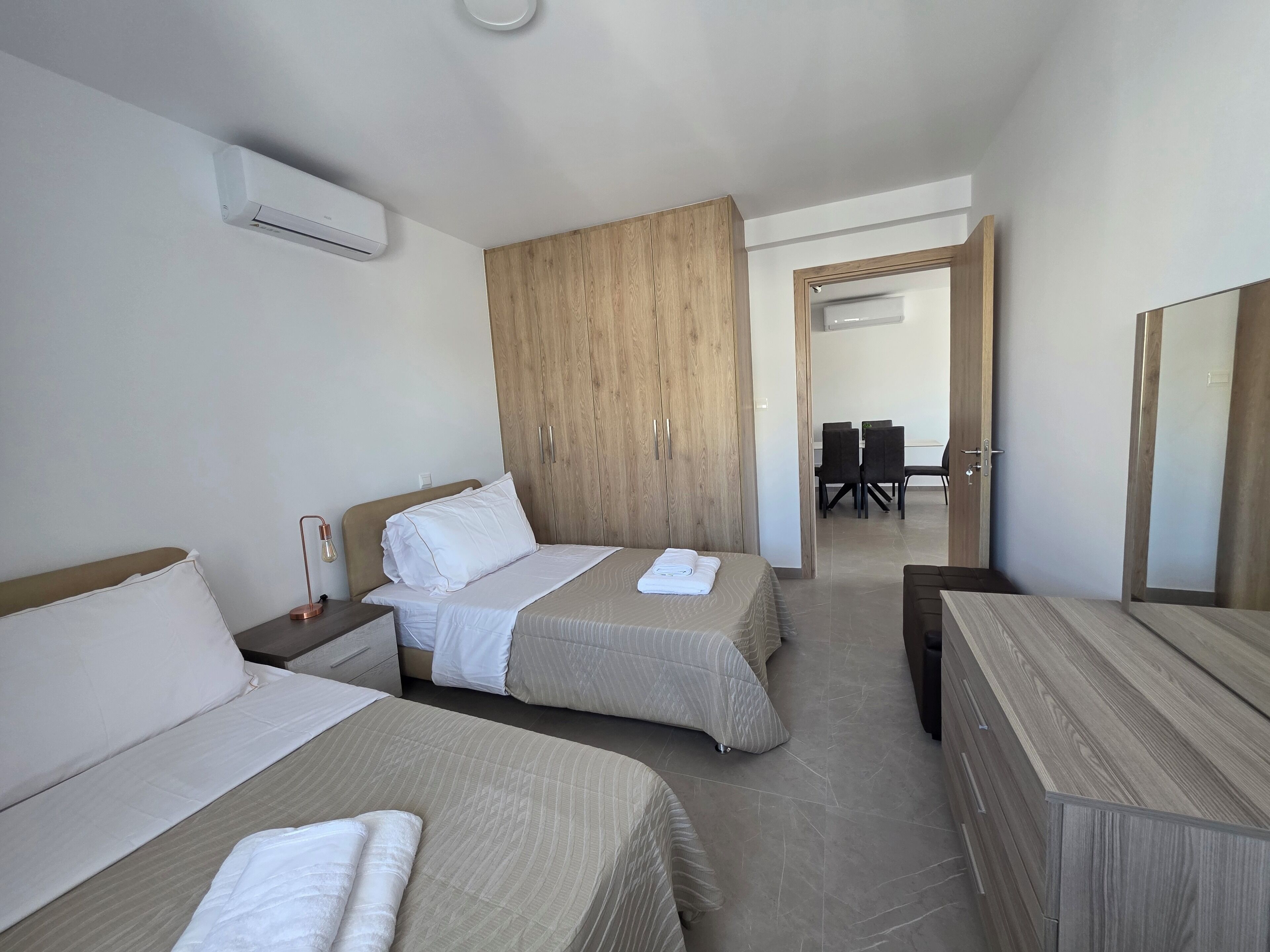 Une chambre d’hôtel avec deux lits, une armoire en bois, un bureau et un coin repas visible à travers une porte ouverte.
