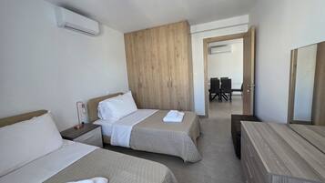 Quarto de hotel com duas camas, um guarda-roupa de madeira, uma mesa de escritório e uma área de refeições visível através de uma porta aberta.