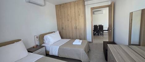 A hotel room with two beds, a wooden wardrobe, a desk, and a dining area visible through an open door.