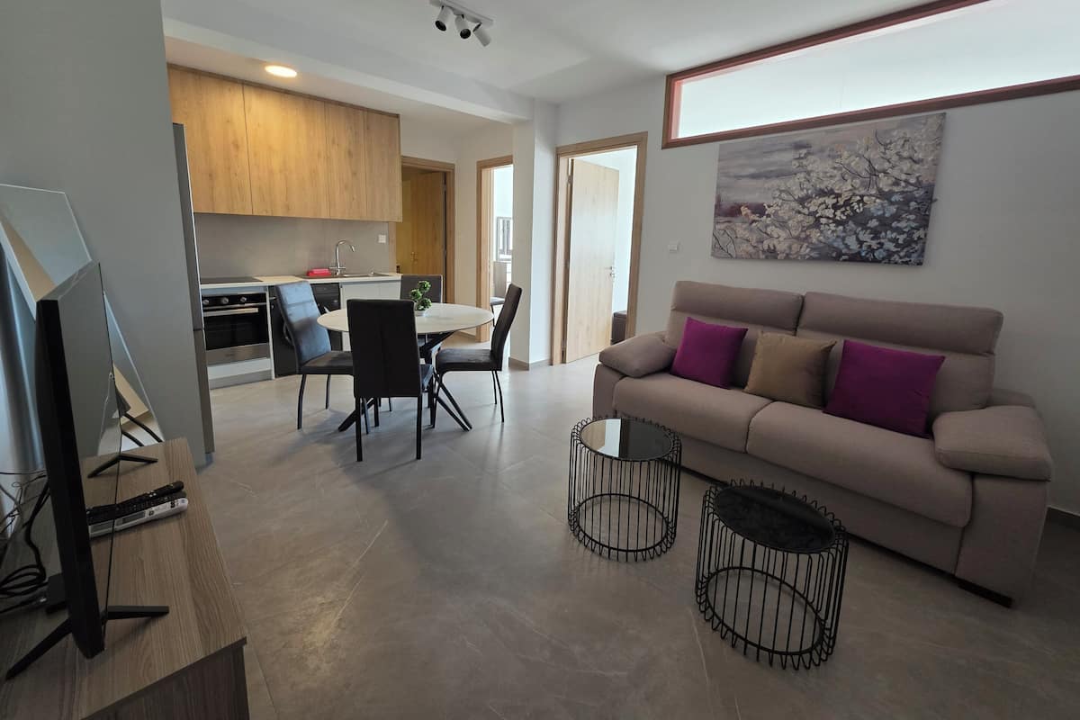 Ein modernes Wohnzimmer mit einem beigefarbenen Sofa, einem Essbereich mit Tisch und Stühlen, einer Küche mit Holzausbau und einem Bild an der Wand.