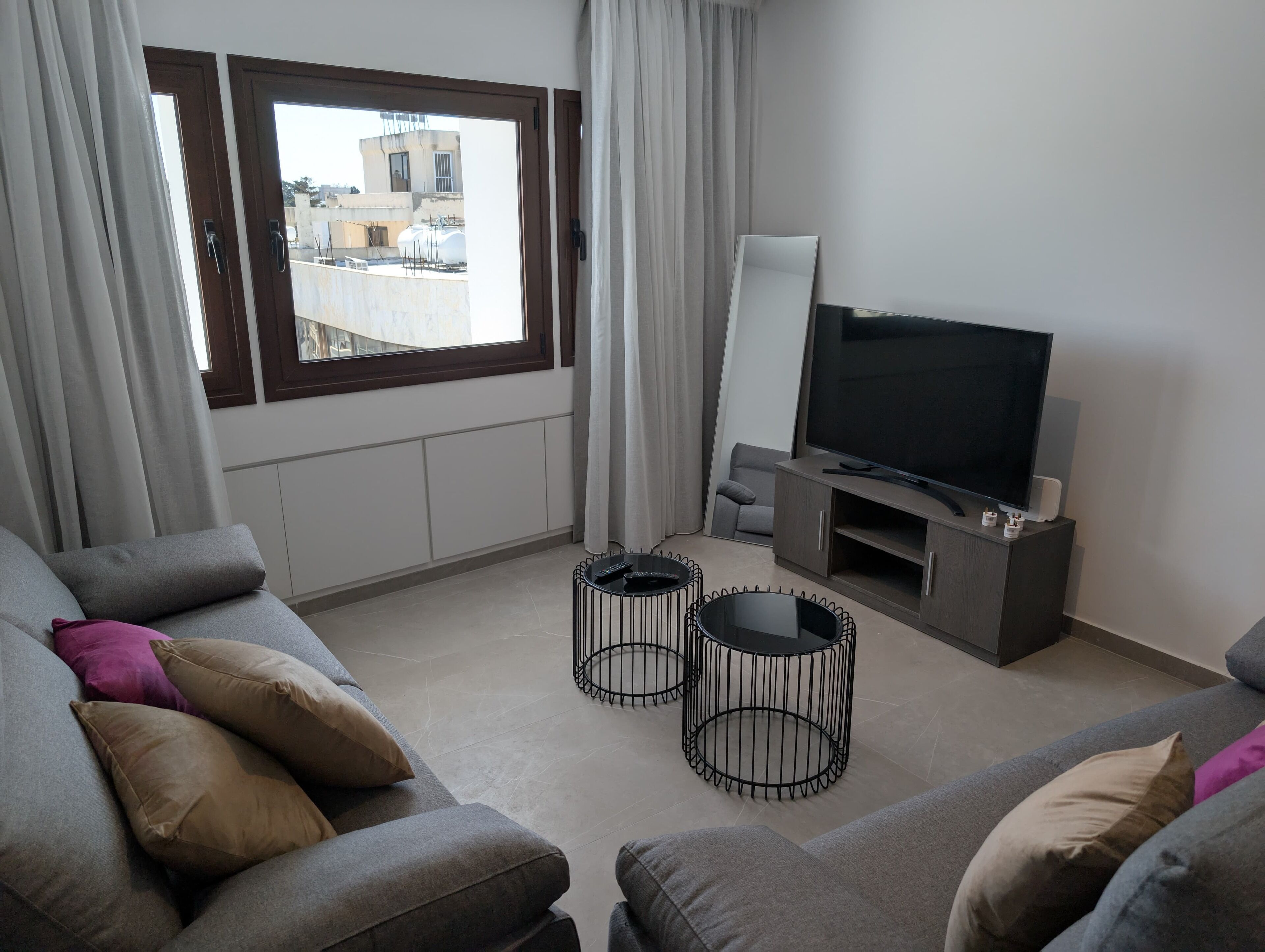 A modern living room with a grey sofa, two black coffee tables, a flat-screen TV on a wooden stand, and a view of buildings through a large window.