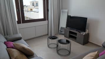 A modern living room with a grey sofa, two black coffee tables, a flat-screen TV on a wooden stand, and a view of buildings through a large window.