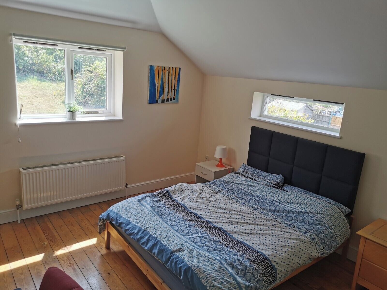 A bedroom with a large bed, two windows, a radiator, a bedside table with a lamp, and a painting on the wall.