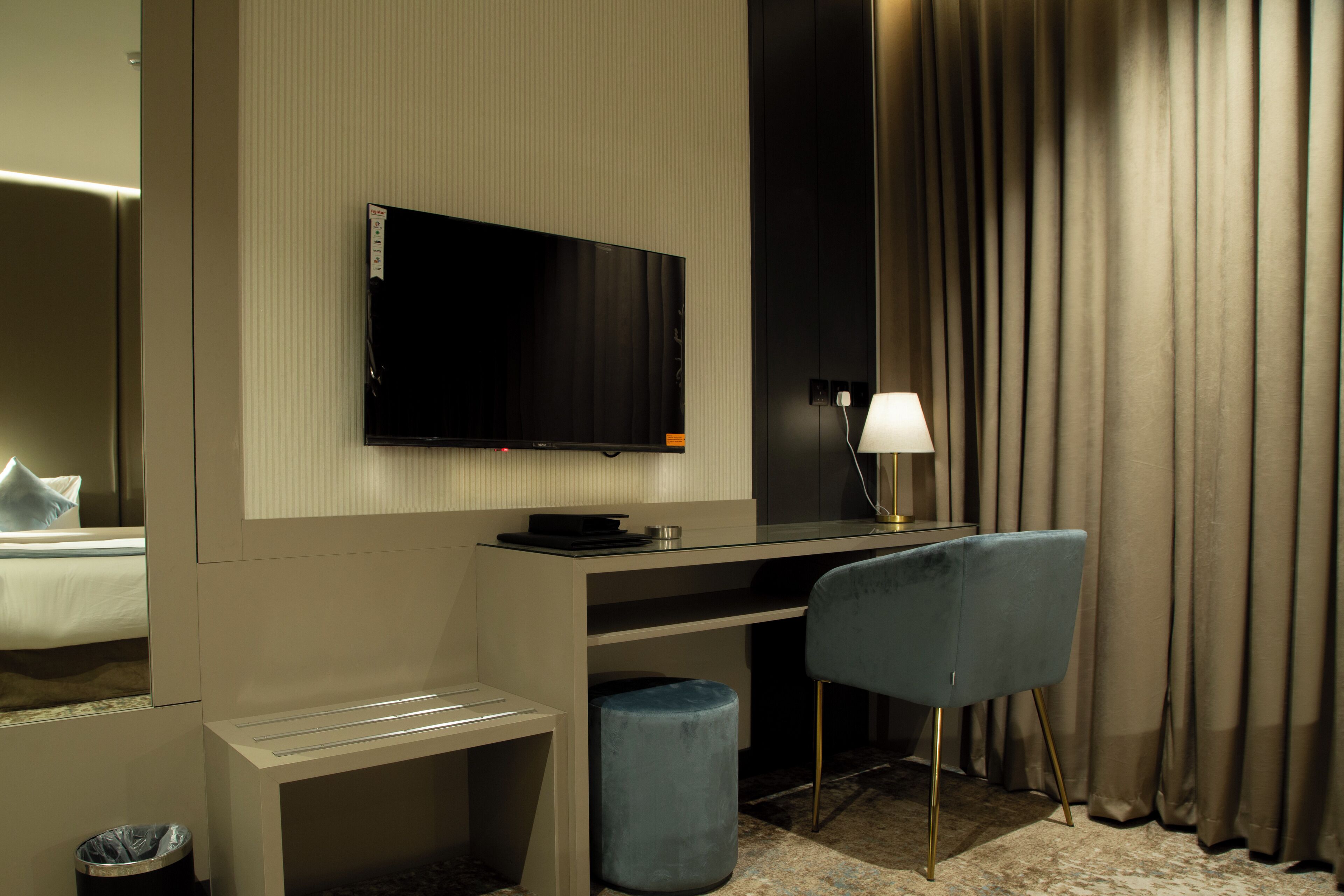 A modern hotel room with a flat-screen TV mounted on the wall, a desk with a chair, a small trash bin, and a lamp.