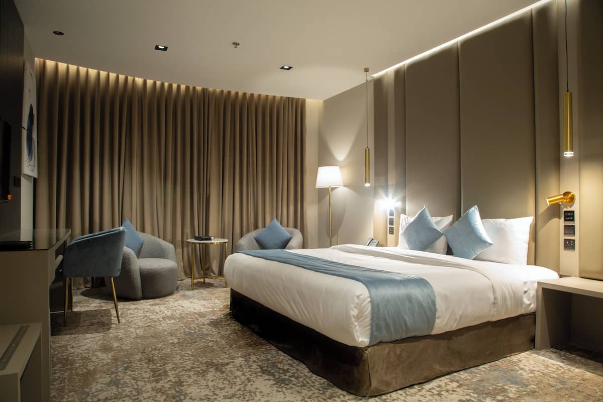 A modern hotel room with a large bed, a seating area with a chair and a small table, and a large window with curtains.