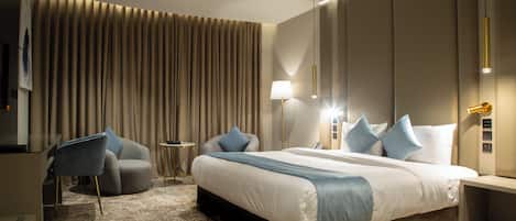 Una habitación de hotel moderna con una cama grande, una zona de estar con una silla y una mesa pequeña, y un ventanal con cortinas.