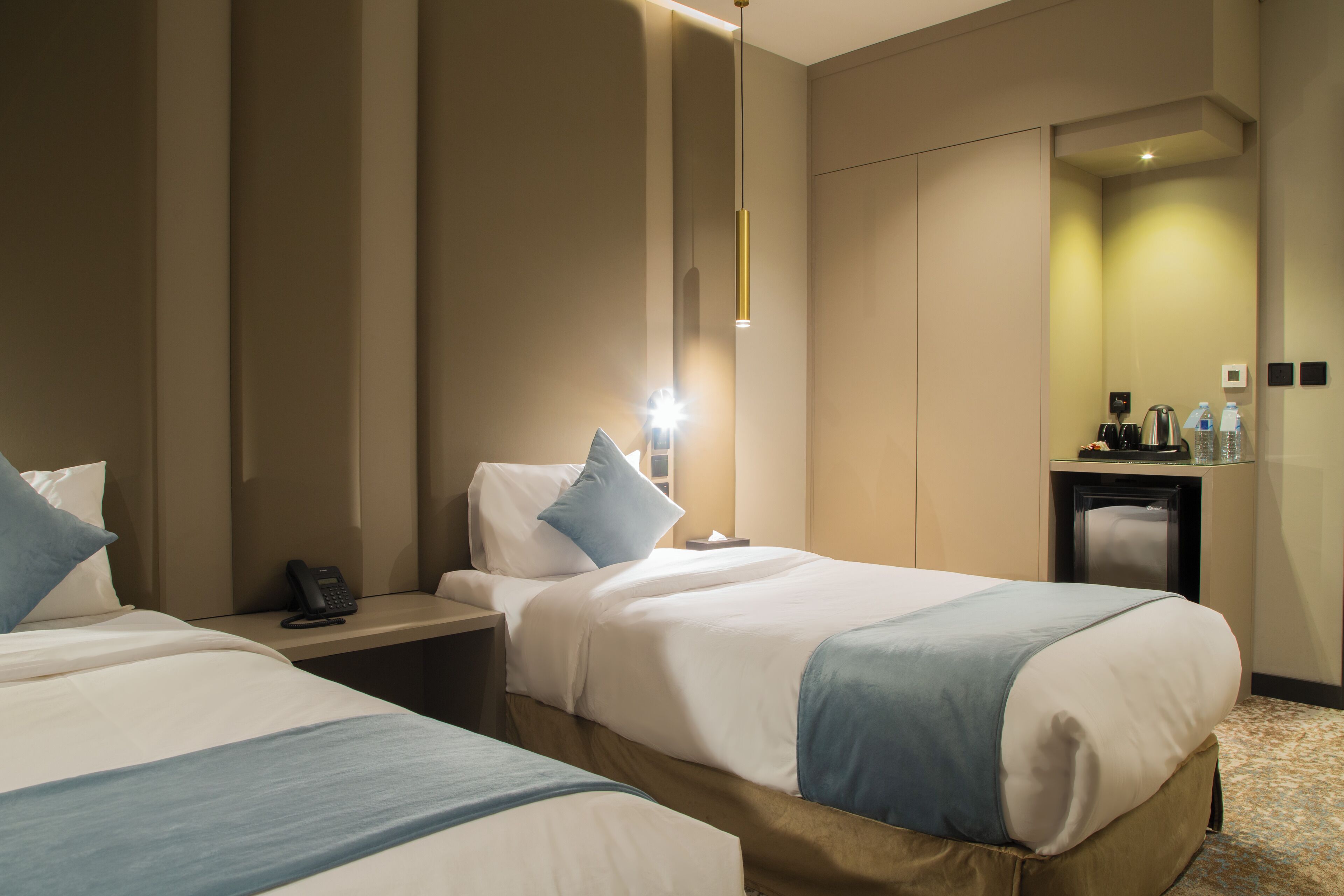 A hotel room with two beds, a bedside table with a phone, a nightstand, a lamp, a small table with a kettle, and a mirror on the wall.