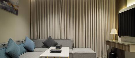 A modern hotel room with a grey sofa, a small white table, a TV, and a large window with curtains.
