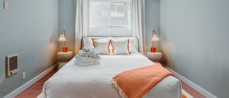 A neatly made bed with white linens and a folded towel stack, two bedside lamps, a window with white curtains, and an air conditioning unit on the wall.