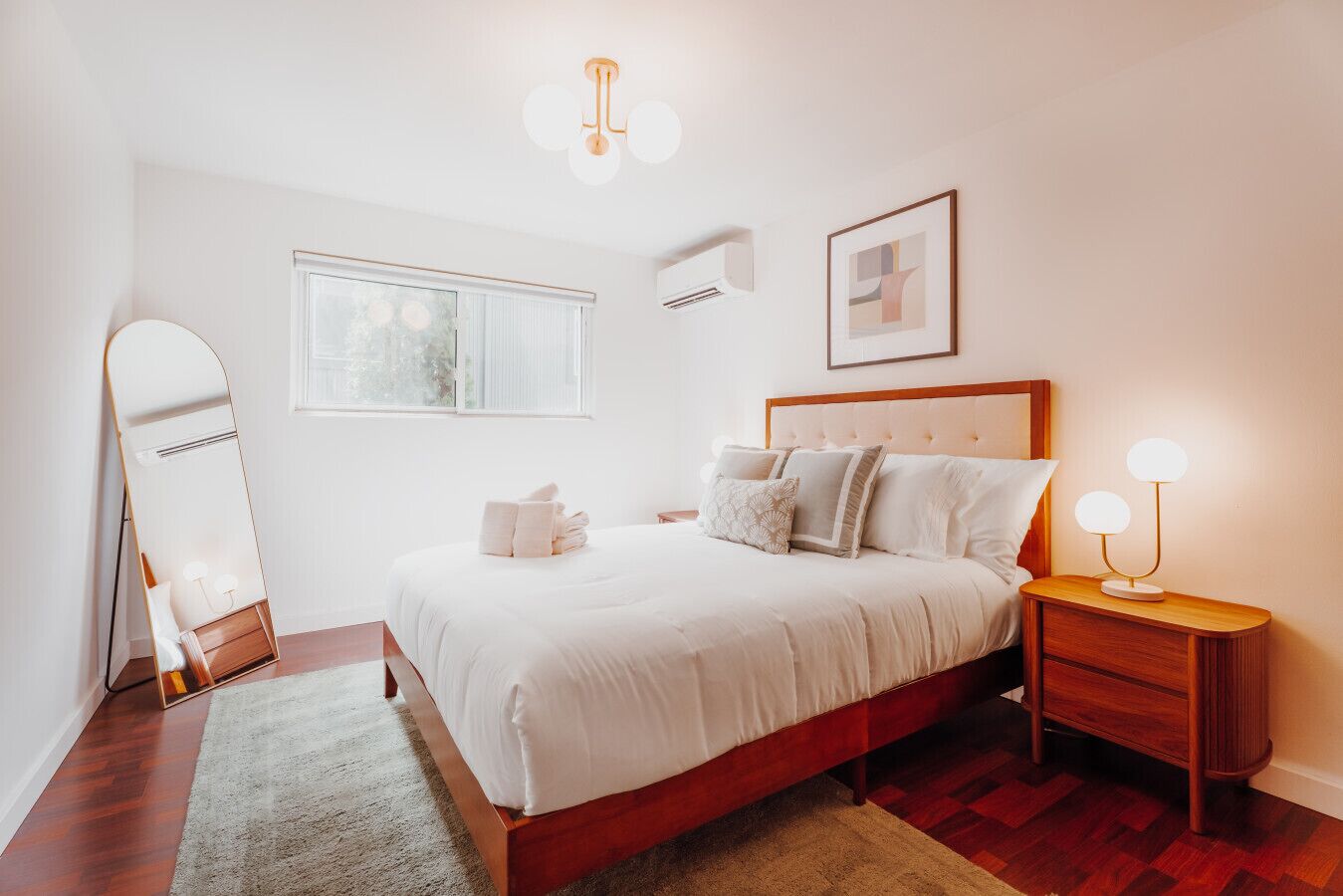 A bedroom with a bed, bedside tables, a mirror, a window, and a wall-mounted light fixture.