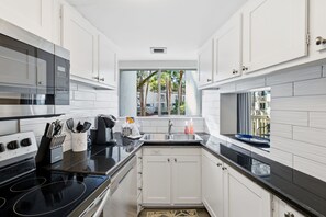 Cozinha moderna com armários brancos, bancada preta, eletrodomésticos de aço inoxidável e janela com vista para palmeiras.