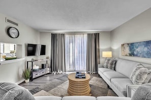 A modern living room with a sofa, a coffee table, a flat-screen TV, a clock, and a large window with curtains.