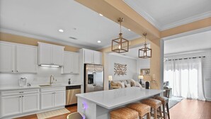 A modern kitchen with white cabinetry, a central island with bar stools, and pendant lighting. - Beautiful Reserve Townhouse - FLETC Welcome! (Saint Simons Island)
