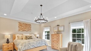A bedroom with a large bed, bedside tables, a dresser, a chair, and a chandelier. - Beautiful Reserve Townhouse - FLETC Welcome! (Saint Simons Island)