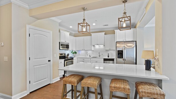A modern kitchen with white cabinetry, a large island with bar stools, pendant lighting, and stainless steel appliances. - Beautiful Reserve Townhouse - FLETC Welcome! (Saint Simons Island)
