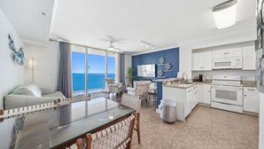 A modern kitchen with white cabinets, a microwave, and an oven. There is a dining area with a glass table and chairs. A TV is mounted on the wall, and a plant is on the counter. - Sept/Oct Open!* Beautiful condo! (Panama City Beach)
