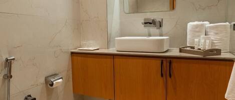 A modern bathroom with a marble wall, a wooden vanity, a white sink, and a shower area.