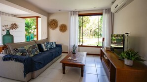 A living room with a sofa, coffee table, and TV. There are two windows with curtains, a wall-mounted air conditioner, and a decorative wall piece. - Bz44 200 metros da praia Área Gourmet (Buzios)