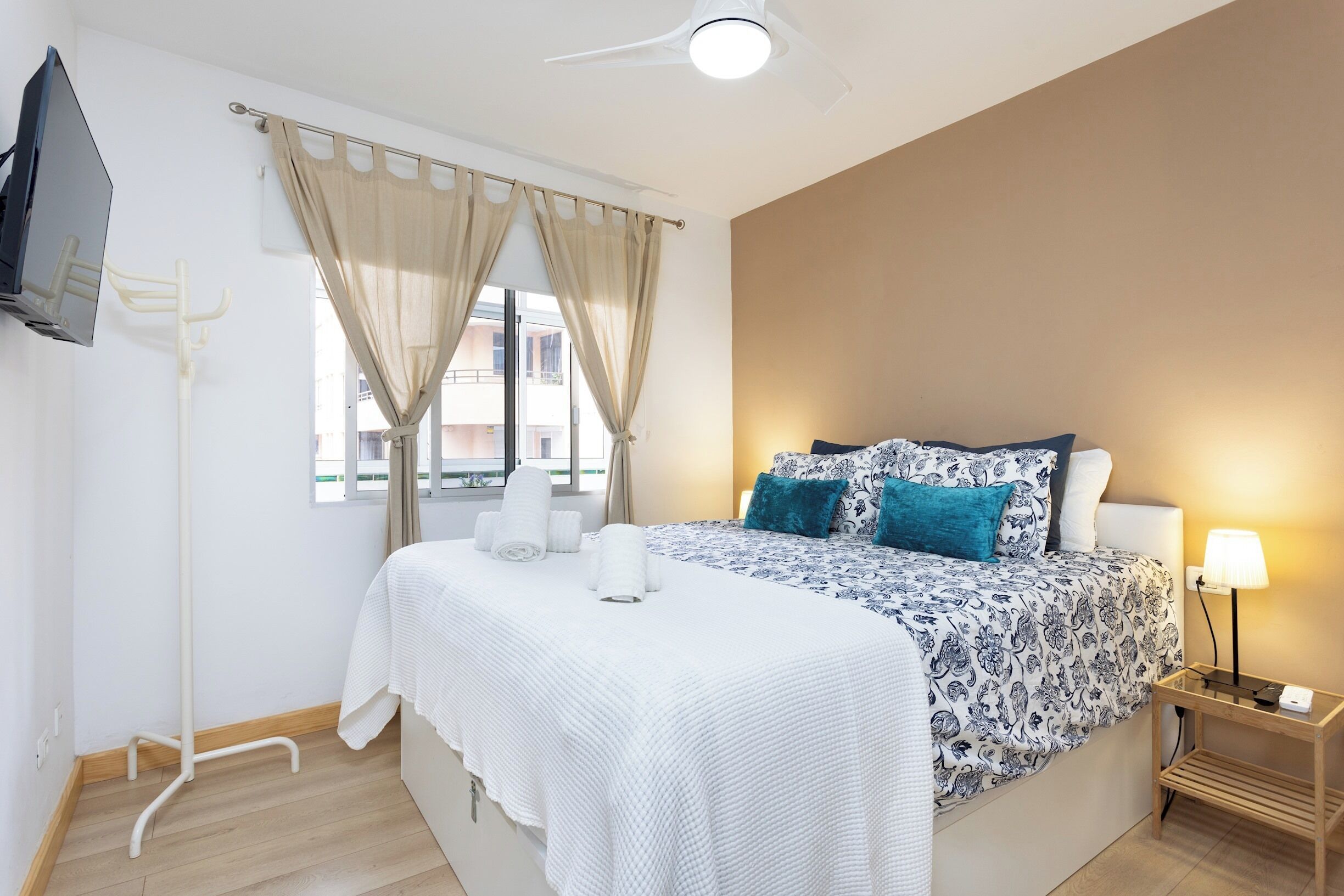 Habitación de hotel ordenada con cama, mesita de noche con lámpara, televisor montado en la pared y ventana con cortinas.