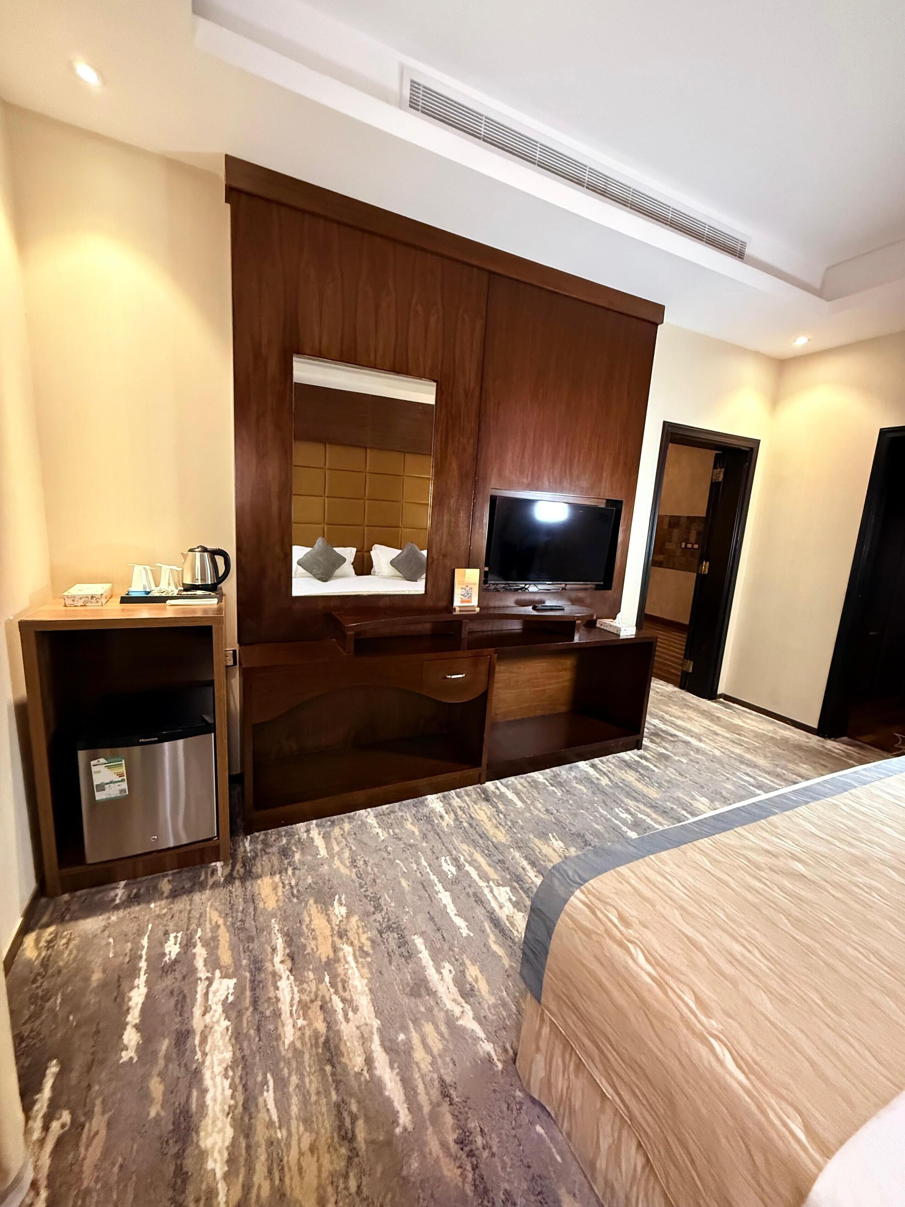 Habitación de hotel con cama, escritorio con televisor de pantalla plana, minibar y espejo grande.