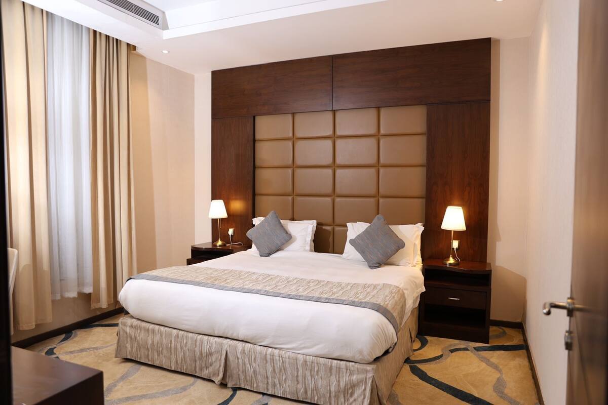 A hotel room with a large bed, two bedside tables with lamps, a wooden headboard, and a patterned carpet.