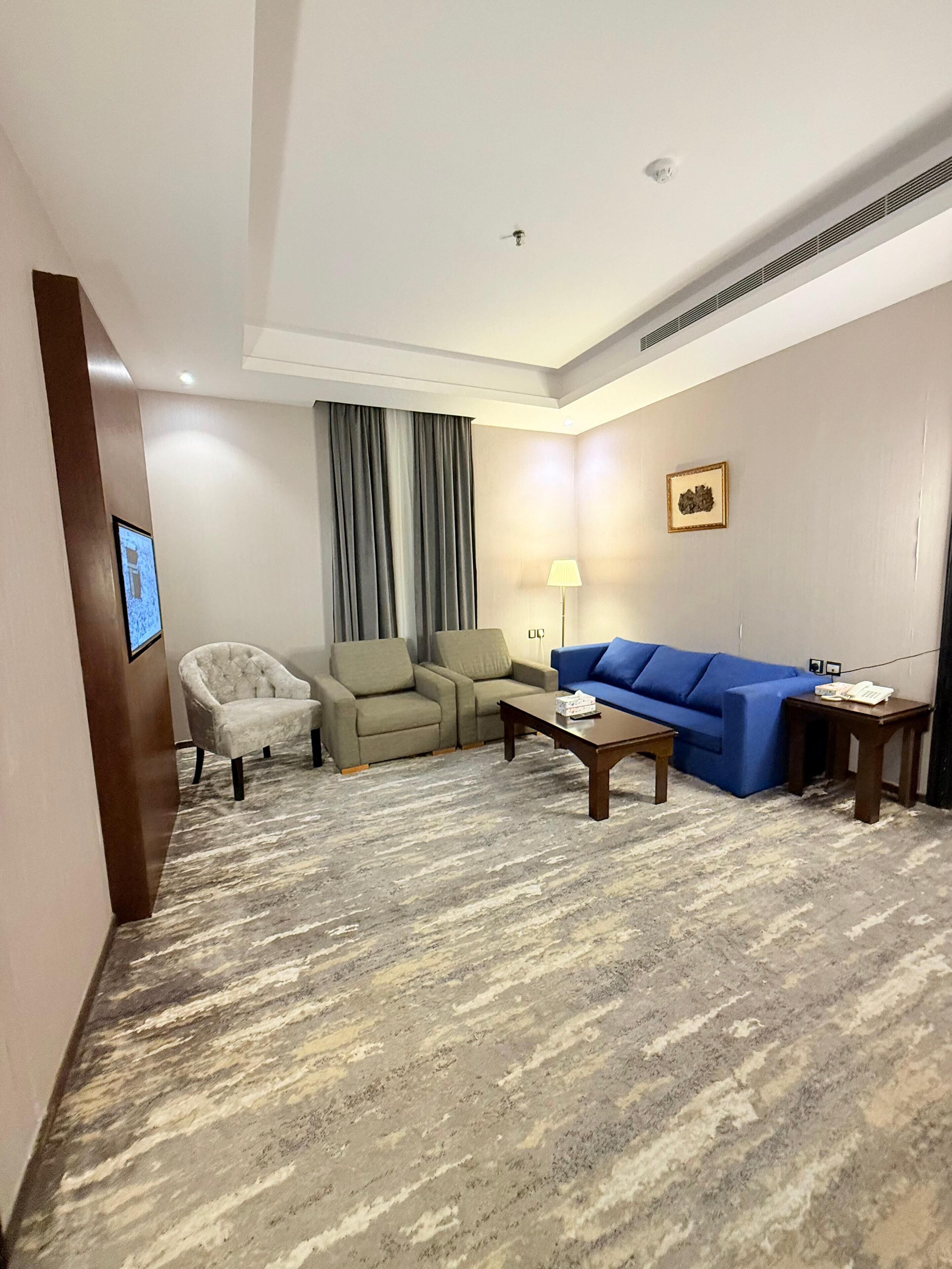A hotel room with a grey carpet, a beige armchair, a blue sofa, a wooden coffee table, and a lamp.