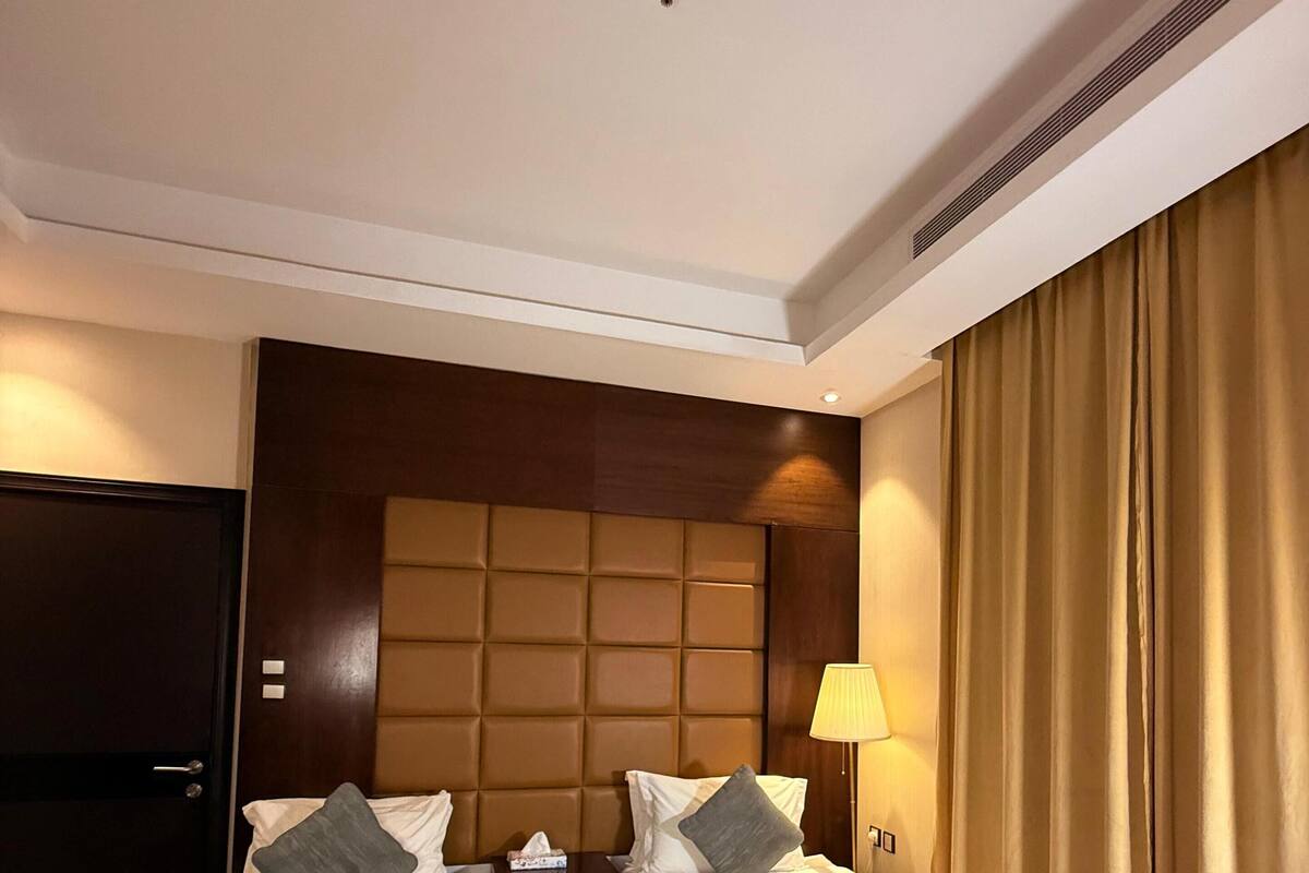 A hotel room with two beds, a wooden headboard, a nightstand, a lamp, and a window with curtains.