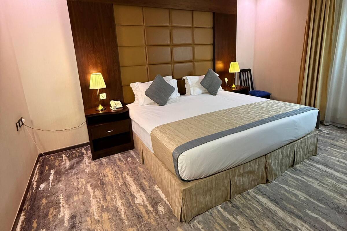 A hotel room with a large bed, a wooden headboard, a nightstand with a lamp, and a carpeted floor.