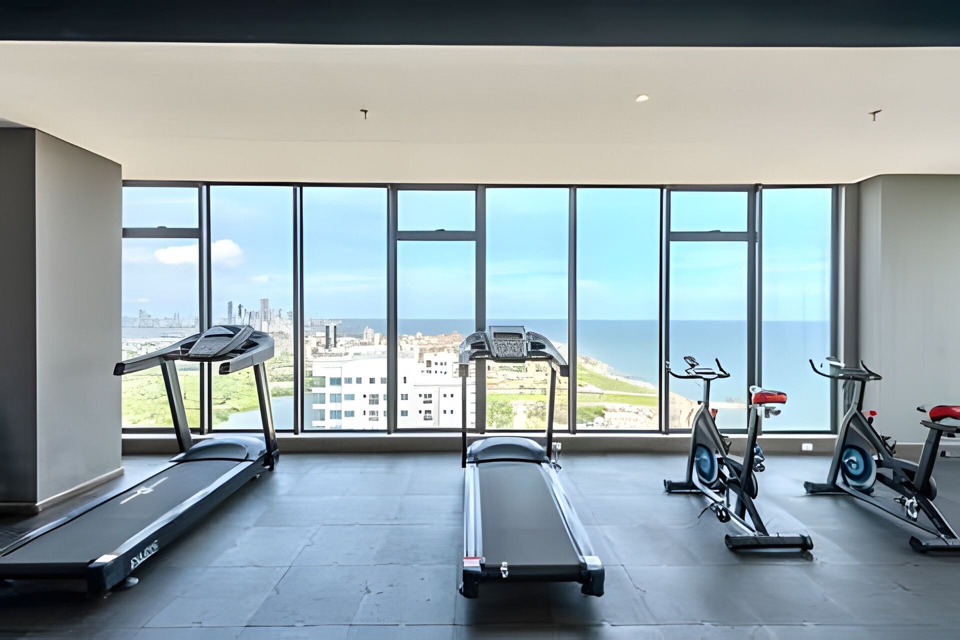 Un gimnasio con cintas de correr, elípticas y bicicletas estáticas, con grandes ventanales que ofrecen vistas a la ciudad y al océano.