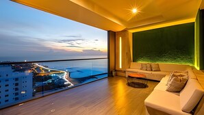 Una sala de estar moderna con un balcón grande, un sofá y vistas a una ciudad costera al atardecer.