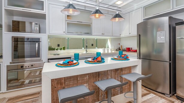 Una cocina moderna con electrodomésticos de acero inoxidable, una isla central con taburetes de bar, iluminación colgante y un espejo grande sobre el lavamanos.