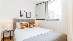 A modern hotel room with a large bed, two bedside tables with lamps, a grey upholstered headboard, and a large window with a view of buildings. - Infante Studio w/ Parking by LovelyStay (Vila Nova de Gaia)