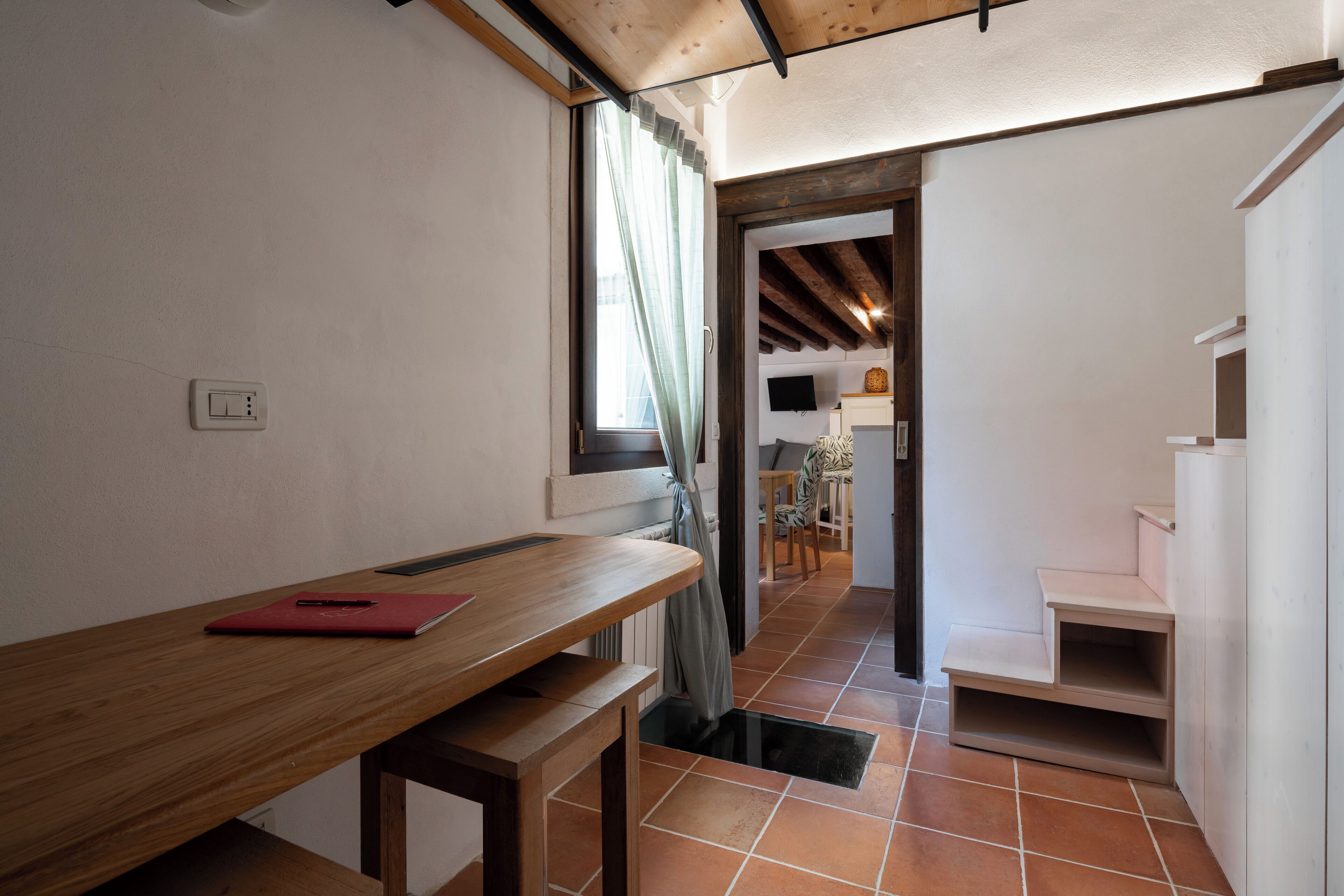 A narrow hallway with a wooden bench, a red notebook, and a window with curtains.